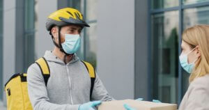 Young male courier in medical mask bringing carton boxes to woman on street and tapping on phone to check parcel. Food delivery service concept - What is the Most Affordable Meal Delivery Service
