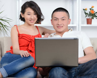 Portrait of young Asian couple sitting on sofa at home, using laptop computer, smiling and learning how to budget for a month