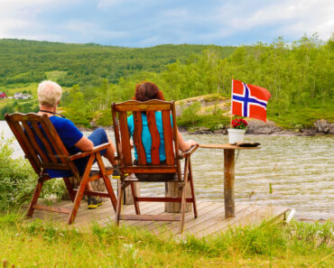 Mature couple woman and man relaxing outdoor, sitting on wooden chairs on lake fjord shore. Holidays relaxation and trip. Norway Scandinavia Europe -