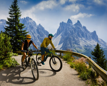 Couple mountain bike riders on electric bike, e-mountainbike rides up mountain trail. Woman and Man riding on bike in Dolomites mountains landscape. Cycling e-mtb enduro trail track. Outdoor sport activity.